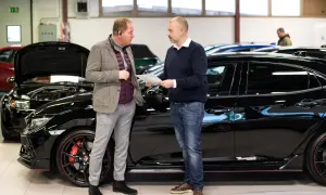 Två män diskuterar papper vid en svart sportbil. De verkar prata om bilkostnad per månad.