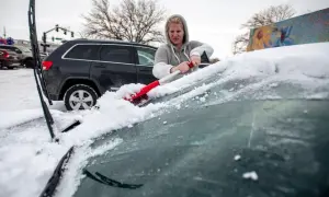 Kvinna skrapar snö från bilrutan med en röd borste. Snön ligger tjock på bilens framruta.
