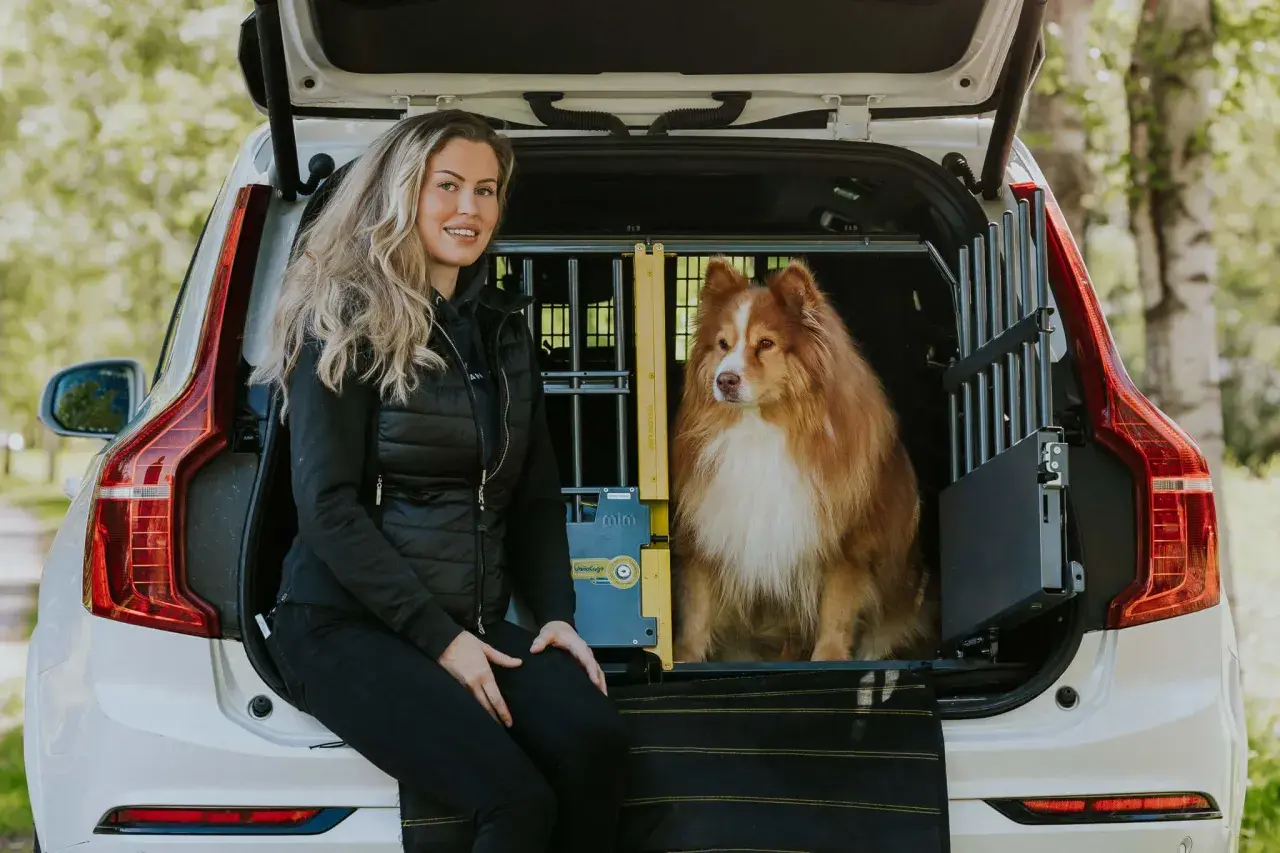En kvinna sitter bredvid en hund i en bilbur. Hunden, en vacker långhårig blandras, tittar ut från buren.