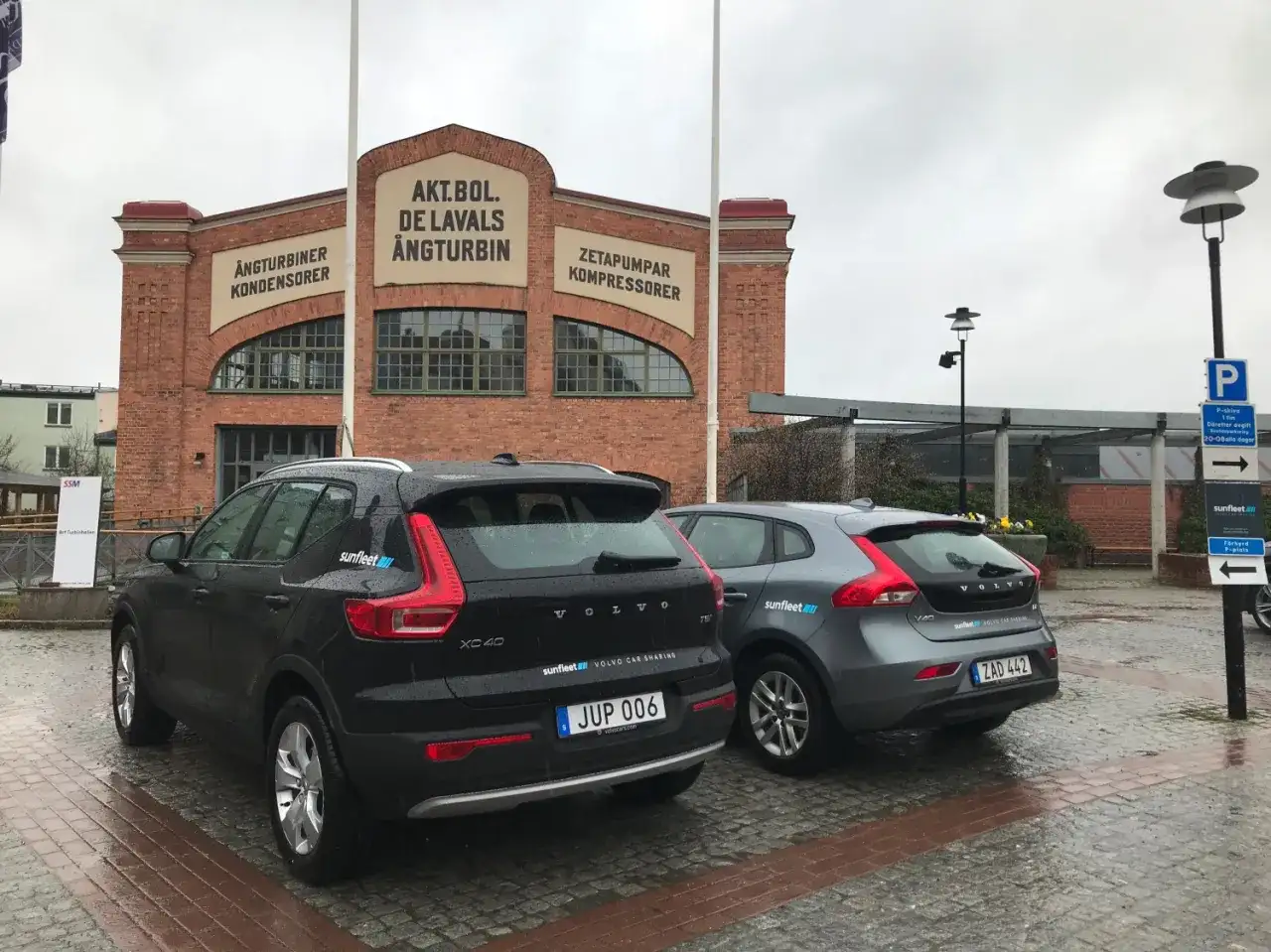 Två Volvo bilar från Volvo on demand står parkerade framför en gammal tegelbyggnad med texten "AKT.BOL. DE LAVALS ÅNGTURBIN".