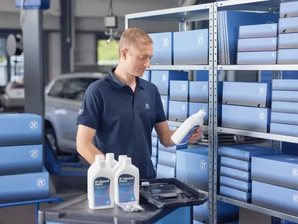 Mekaniker håller en flaska ZF Lifeguard Fluid 8, perfekt för att byta olja i automatlåda. Hyllor fyllda med ZF-produkter.