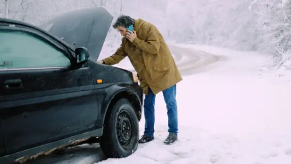 Man med telefon vid en bil med öppen motorhuv. Kallstart bil i snöväder.